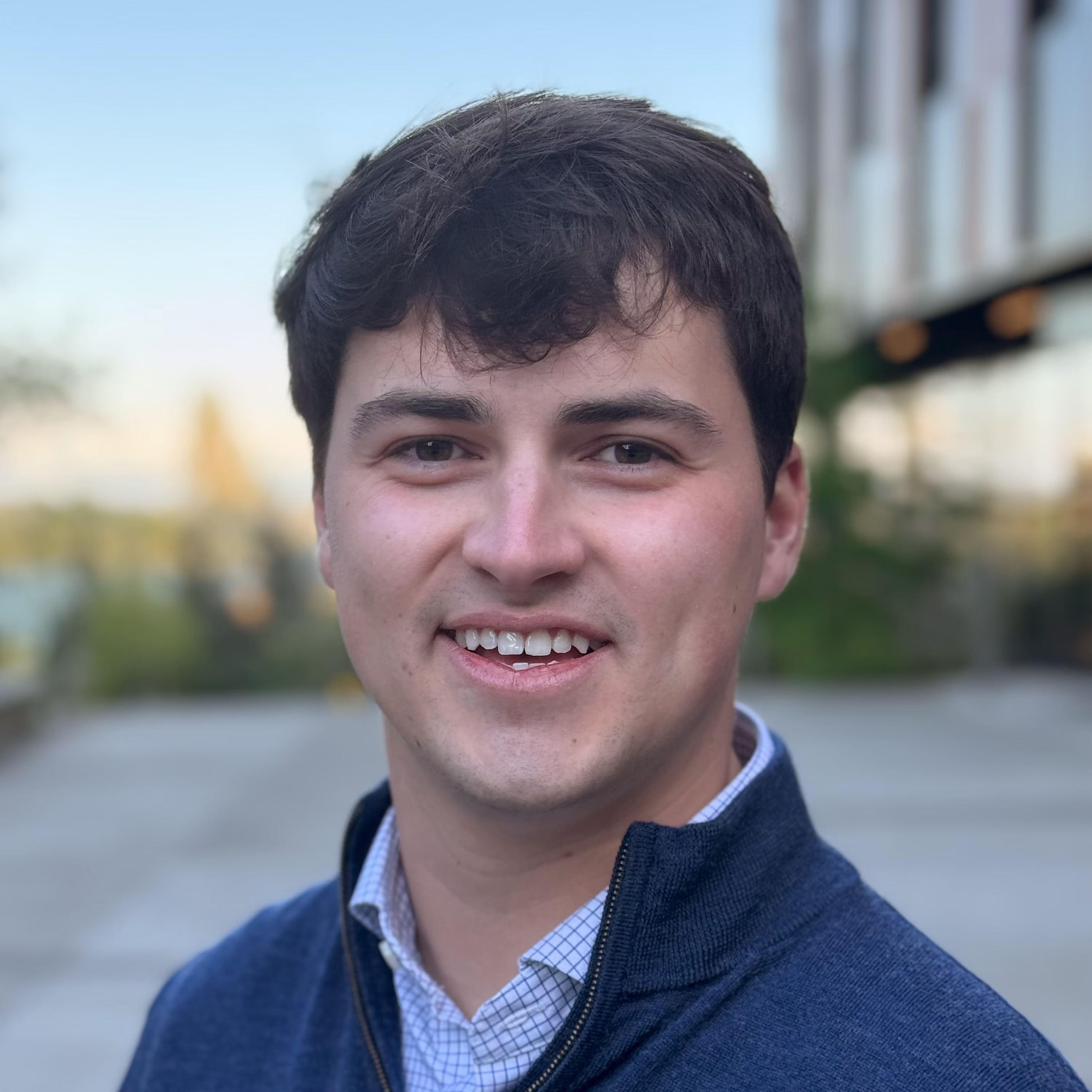 Headshot of James, a 25-year old white man with short brown hair, wearing a collared shirt and blue sweater smiling in front of a blurred outside background.
